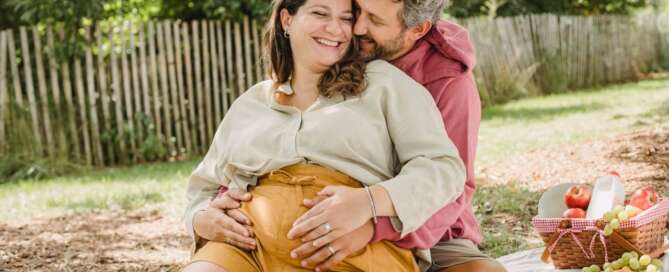 Image shows one person wrapping arms around pregnant person. Your clients can still have safe, kinky fun during pregnancy.