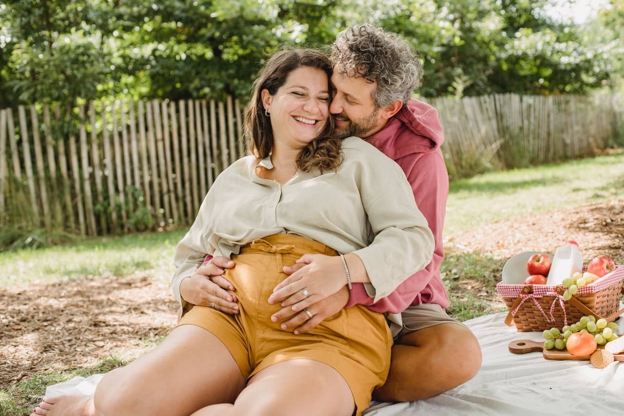 Image shows one person wrapping arms around pregnant person. Your clients can still have safe, kinky fun during pregnancy.