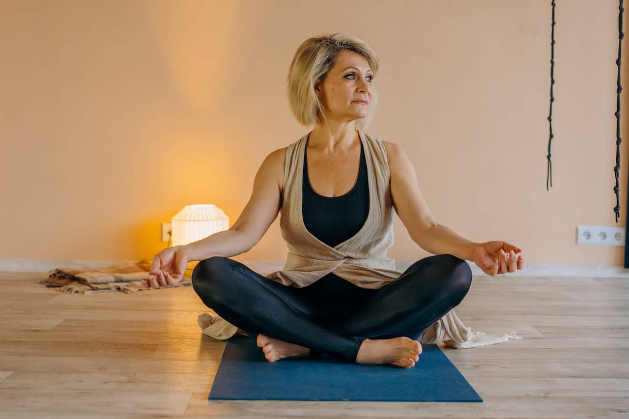 a midlife woman sits on a yoga mat in a cross legged position looking off to the side.