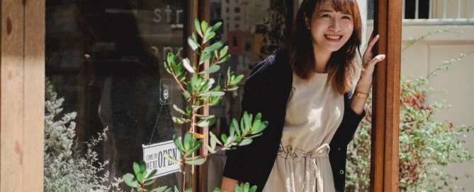 an asian woman wearing an apron who works as a sex toy retailer walks out of her store smiling