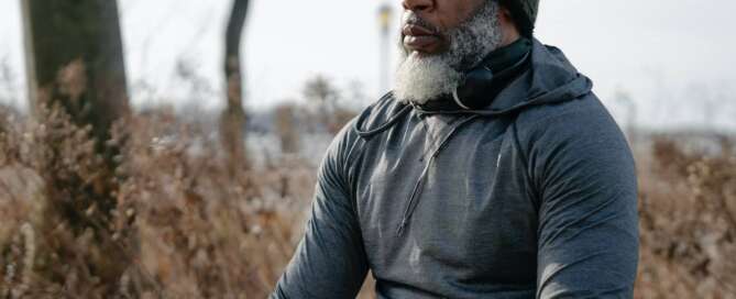A man with a gray beard wearing a hoodie sits outside with his eyes closed practicing self-care