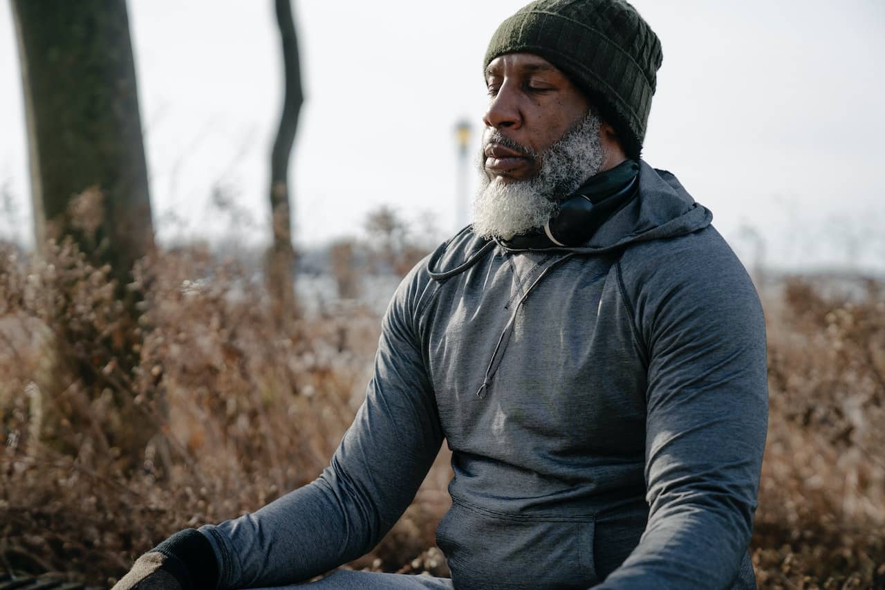 A man with a gray beard wearing a hoodie sits outside with his eyes closed practicing self-care
