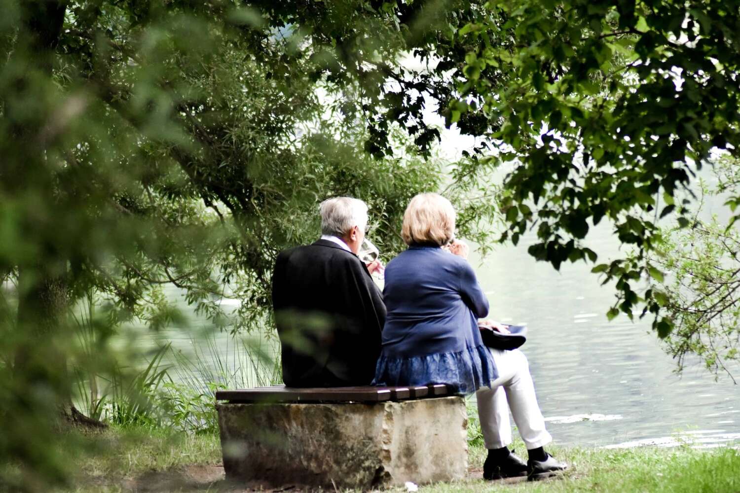 an older couple sits on a bench by the water relaxing with glasses of wine because they have resolved their erectile dysfunction difficulties.