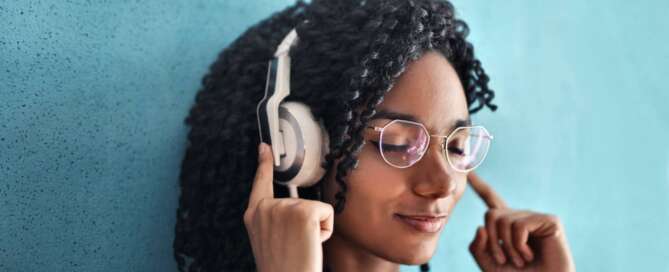 a woman with glasses stands against a blue wall with white headphones on her ears, closing her eyes and practicing her co-active coaching listening skills