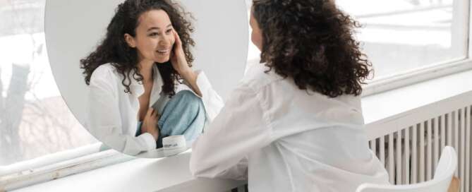 a woman in a white top smiles at herself in the mirror after doing her self-pleasure practice.