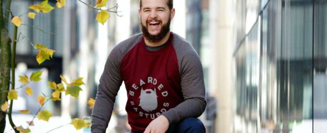 a man in a red and gray shirt sits on a wooden bench smiling because he realizes why people seek sex coaching