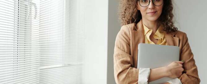 a woman in a beige blazer and glasses hugs a laptop to her body and smiles at the camera after comparing sex coach training programs