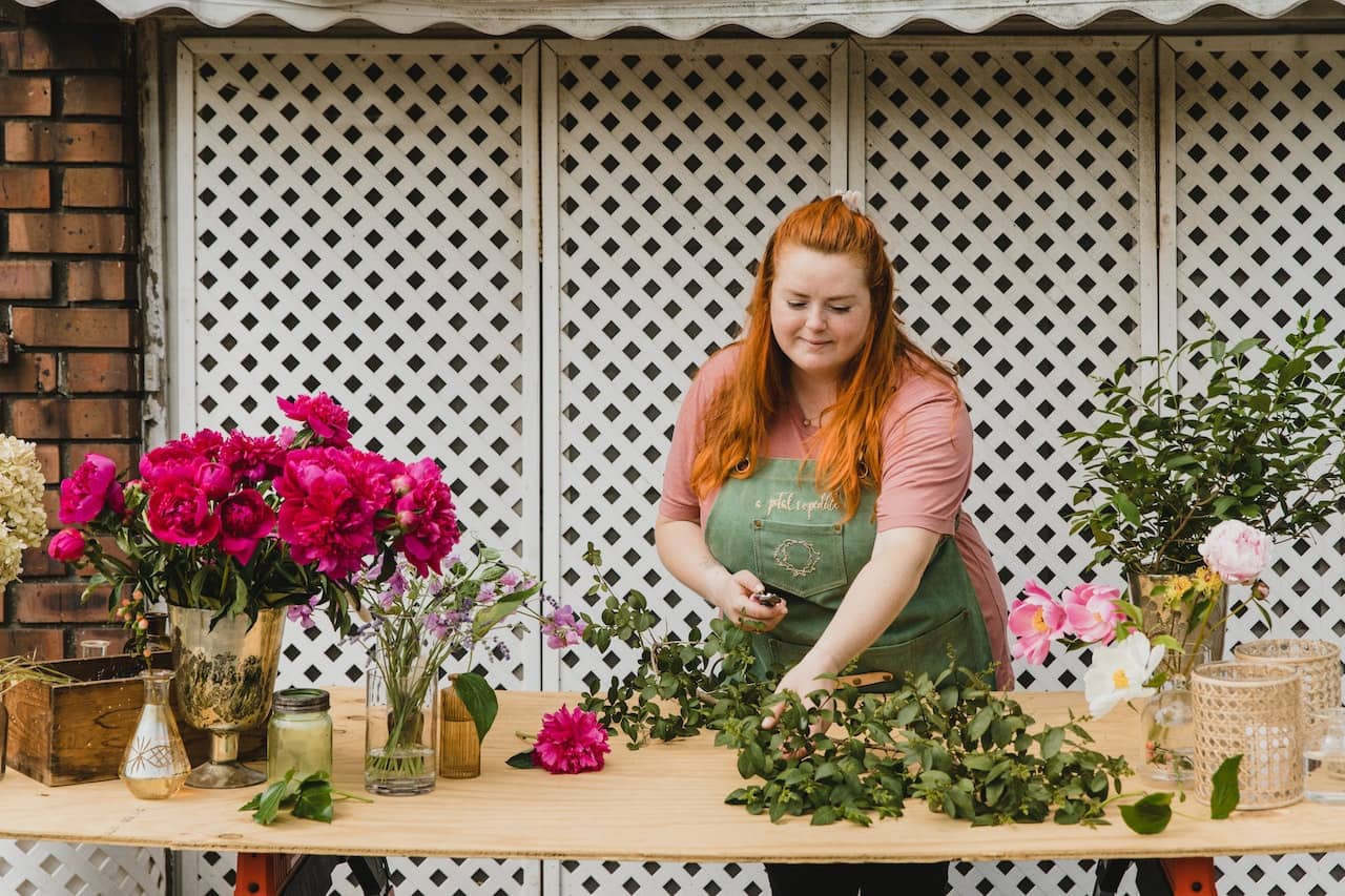 a woman with red hair wearing a green apron stands in front of a wooden table surrounded by flowers, making bouquets after figuring out that she’s ready for a career change.