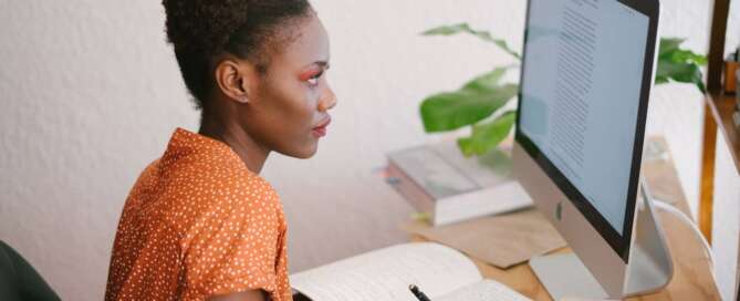 a person wearing an orange shirt with white dots writes in a notebook while looking at a computer desktop after learning how to study as a sex coaching student.