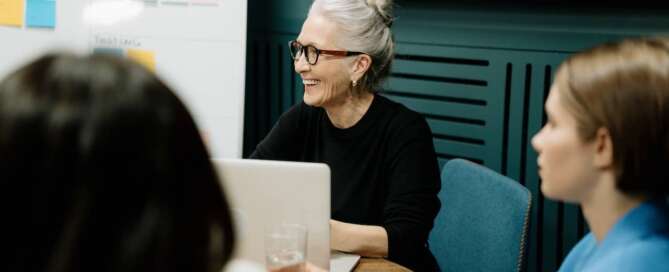 an older woman sits at a round table with a white board behind her, smiling in a direction off camera after transitioning from sex educator to sex coach.