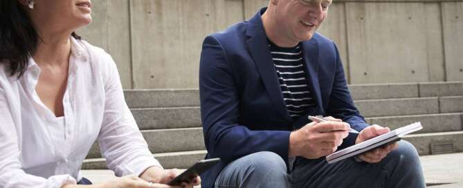 a man in a blazer sits next to a woman in a white shirt smiling at his notepad after learning how to balance his heart-centered work and marketing practice