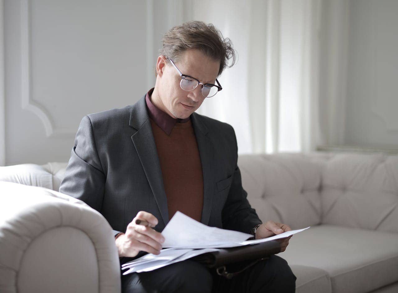 a man wearing a suit jacket and glasses sits on a white coach looking through a stack of papers learning about tiny habits for sex coaching.